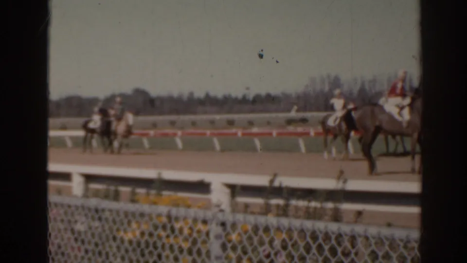 Jockeys and horses move along a Florida racetrack in a soft 8mm frame with railings, fence, and distant trees visible.
