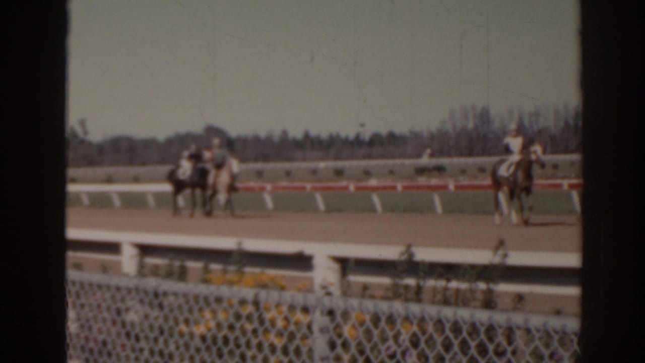 1962 Florida 8mm frame showing horses with riders on a racetrack, viewed from behind a chain-link fence and white rail.