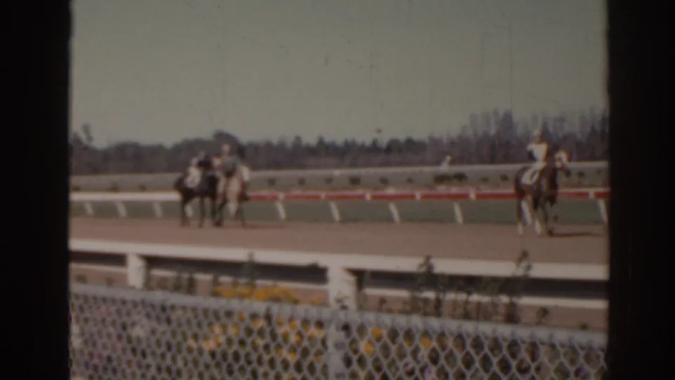 1962 Florida 8mm frame showing horses with riders on a racetrack, viewed from behind a chain-link fence and white rail.