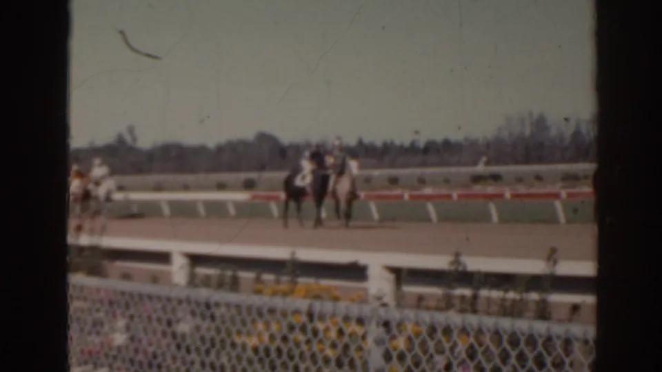 1962 Florida home-movie frame with several horses and riders grouped on the racetrack beyond a foreground fence.