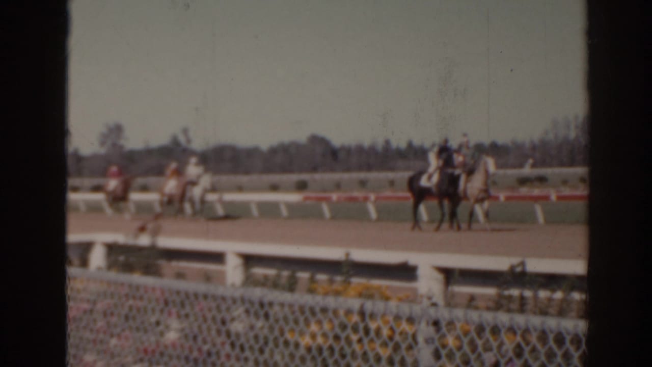 1962 Florida racetrack frame showing riders on horses moving past a white rail, with chain-link fencing close to the camera.
