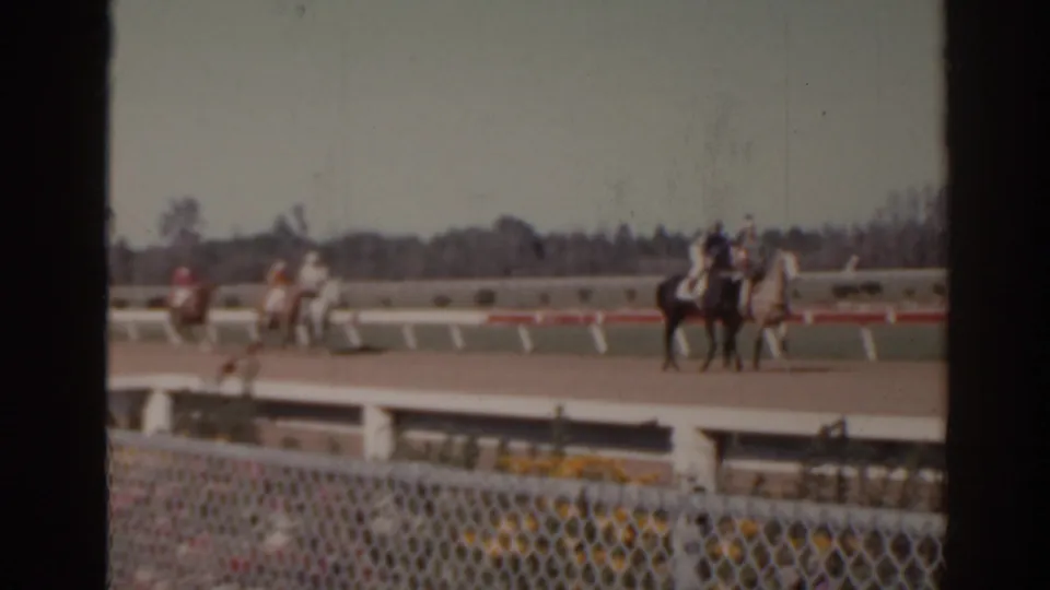 1962 Florida racetrack frame showing riders on horses moving past a white rail, with chain-link fencing close to the camera.