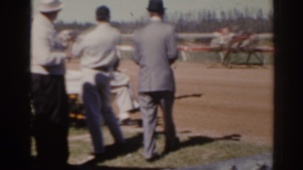 Men in mid-century attire watch horses pass on a Florida dirt racetrack in this 1962 home-movie frame.