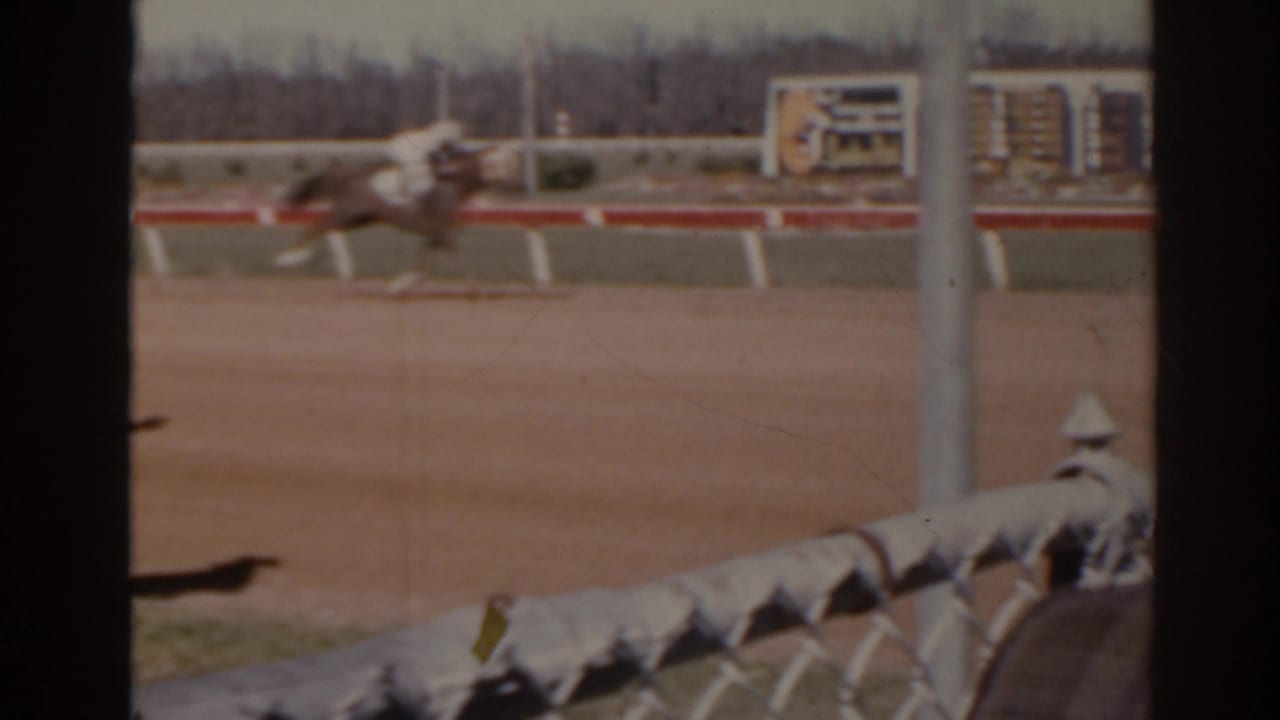 Blurred horse and jockey pass a Florida racetrack rail in a 1962 8mm home-movie frame.