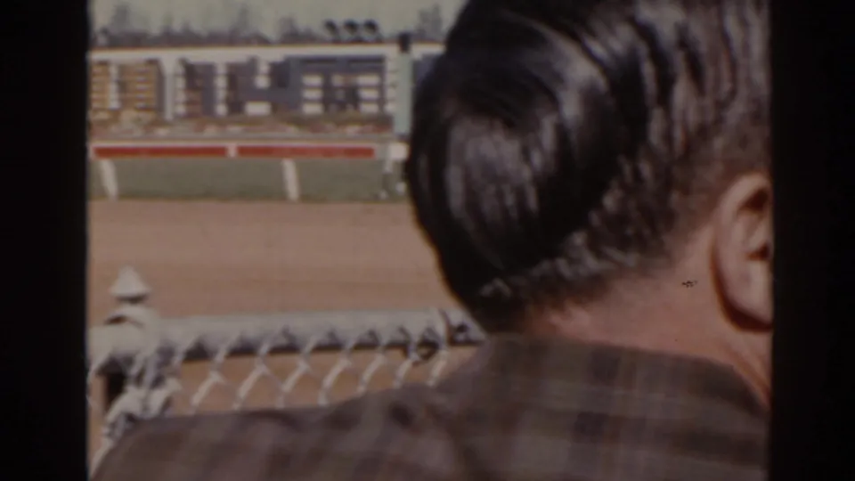 1962 Florida racetrack view with a spectator in a plaid shirt near the camera and the dirt track beyond the fence.