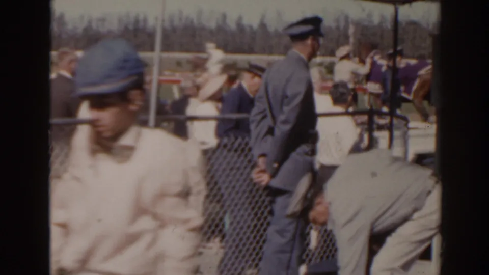 1962 Florida racetrack frame with spectators, uniformed staff, and horses gathered near the chain-link fence.