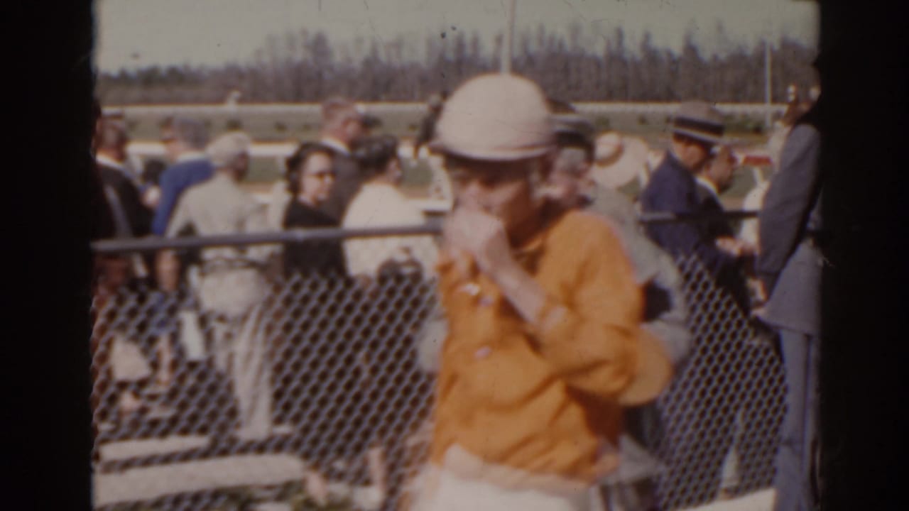 Florida 1962 home-movie frame showing racetrack spectators near a chain-link admission fence as a woman in an orange top passes the camera.