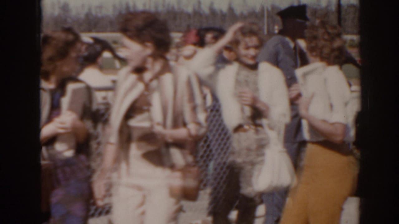 Florida 1962 racetrack crowd frame with women in mid-century dresses and hats gathered near the admission fence.