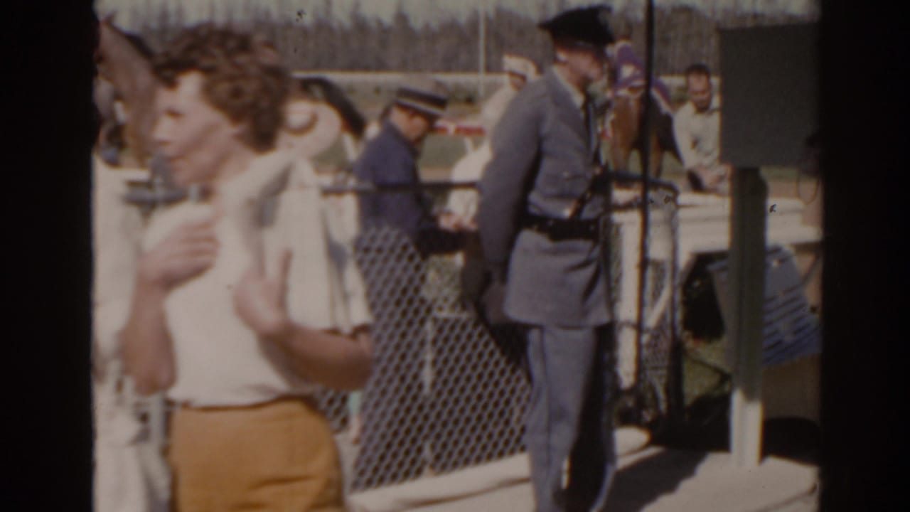 Florida 1962 home-movie frame showing a uniformed official at a racetrack admission gate with spectators moving nearby.