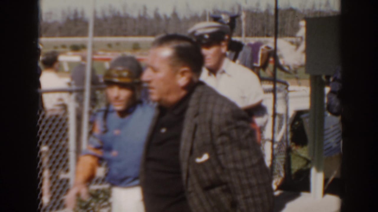 Florida 1962 racetrack frame with a man passing foreground, an official nearby, and a blanketed horse visible behind the fence.