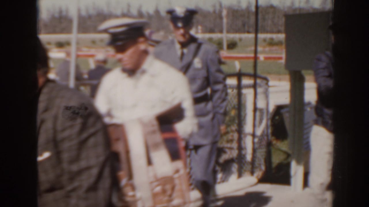 Trackside men near a racetrack gate, including a man in a light shirt and uniformed officials.