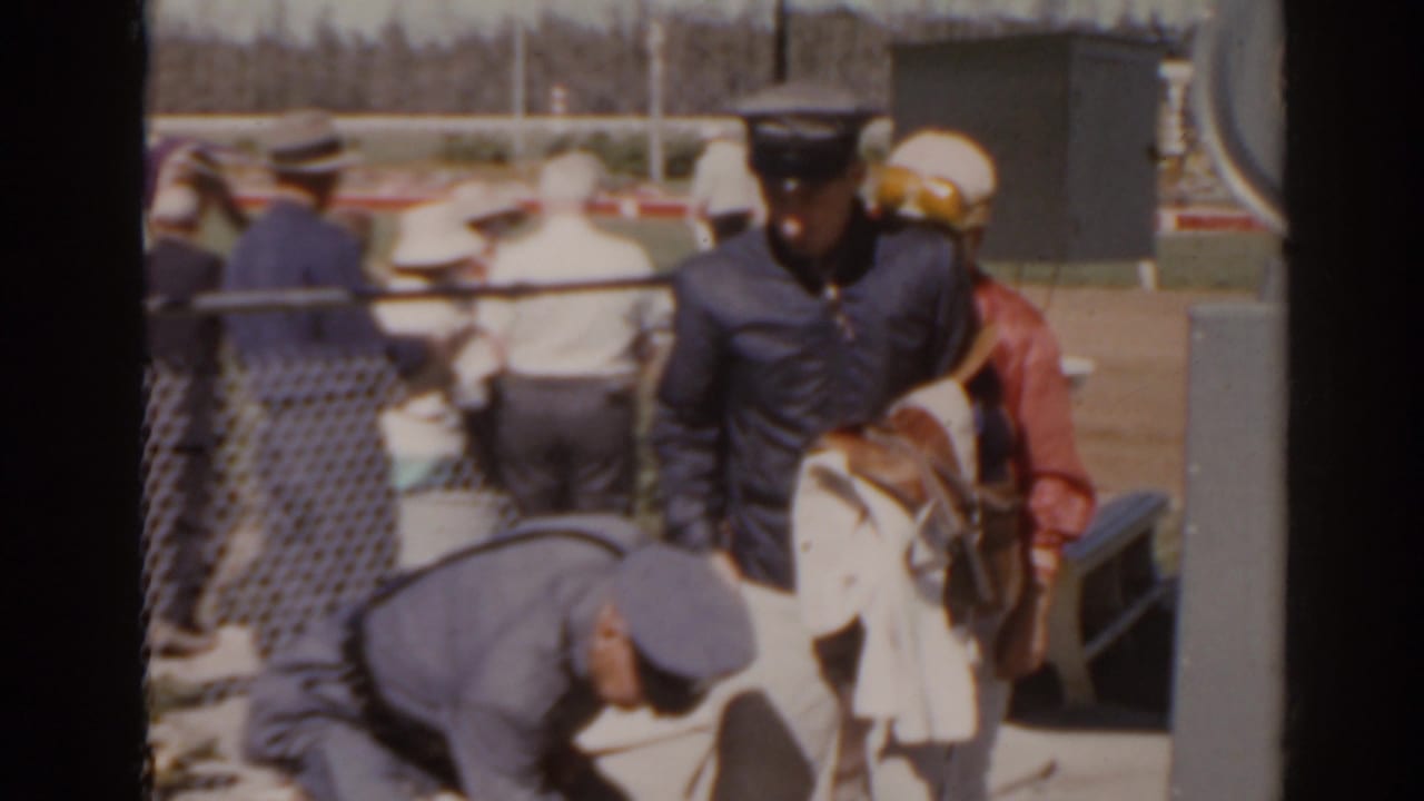 Jockeys and uniformed men stand beside a horse near the starting gate area.
