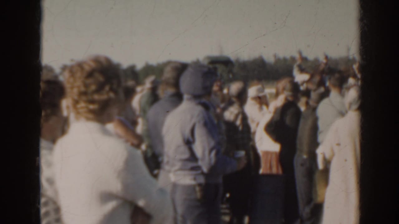 Spectators in 1960s attire gather by the rail at a Florida racetrack during a horse racing event.
