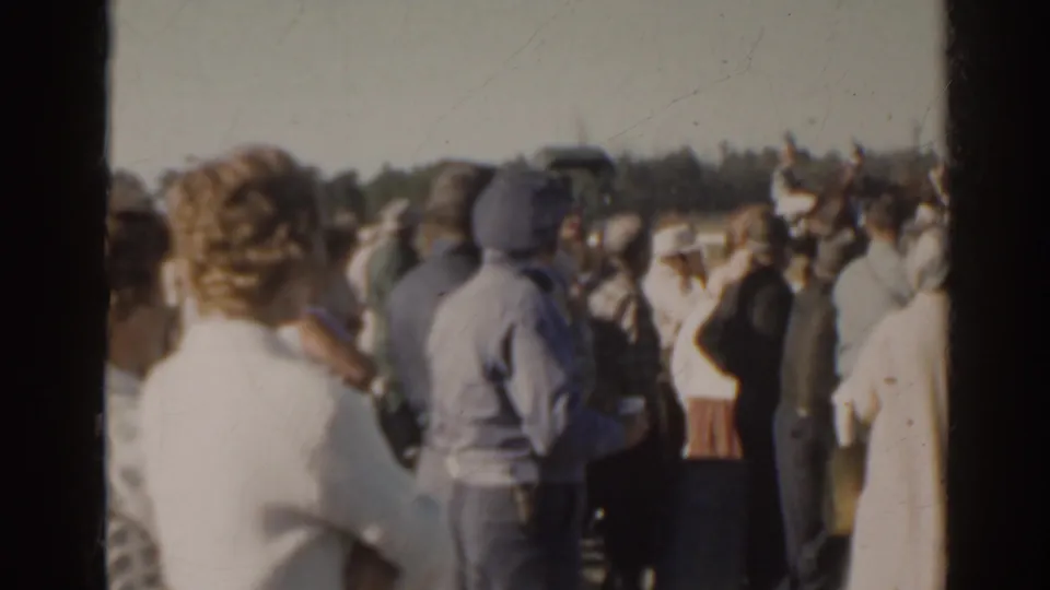 Spectators in 1960s attire gather by the rail at a Florida racetrack during a horse racing event.
