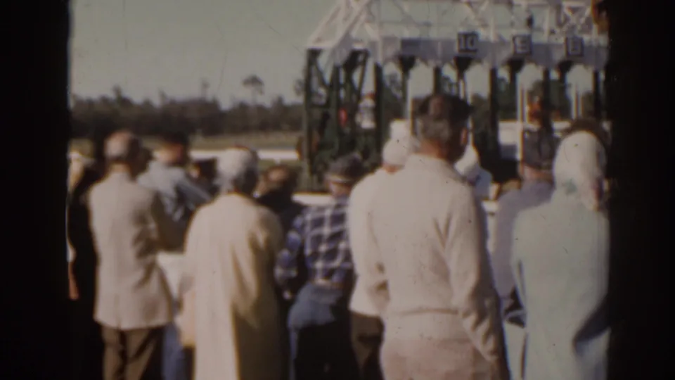 1962 Florida racecourse view with a crowd watching from behind and numbered starting-gate stalls visible across the track.