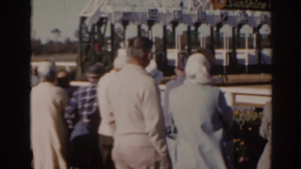 1962 Florida home-movie frame of spectators in light coats watching the racecourse, with starting gates partly visible to the right.