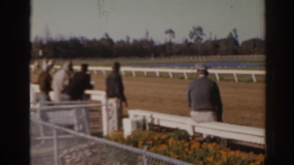 Final 1962 Florida racetrack frame with adults near white rails, flower beds in front, and the racecourse stretching across the background.
