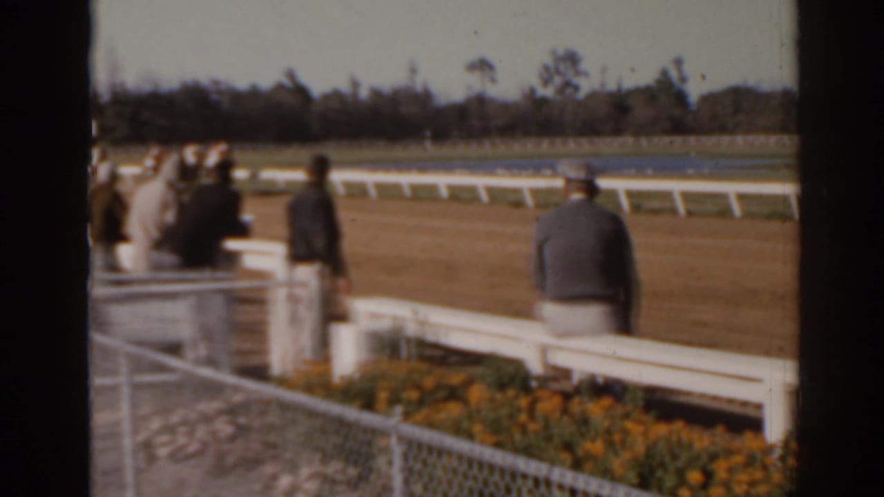 Florida 1962 home-movie frame showing spectators near a white rail as horses race in the distance.