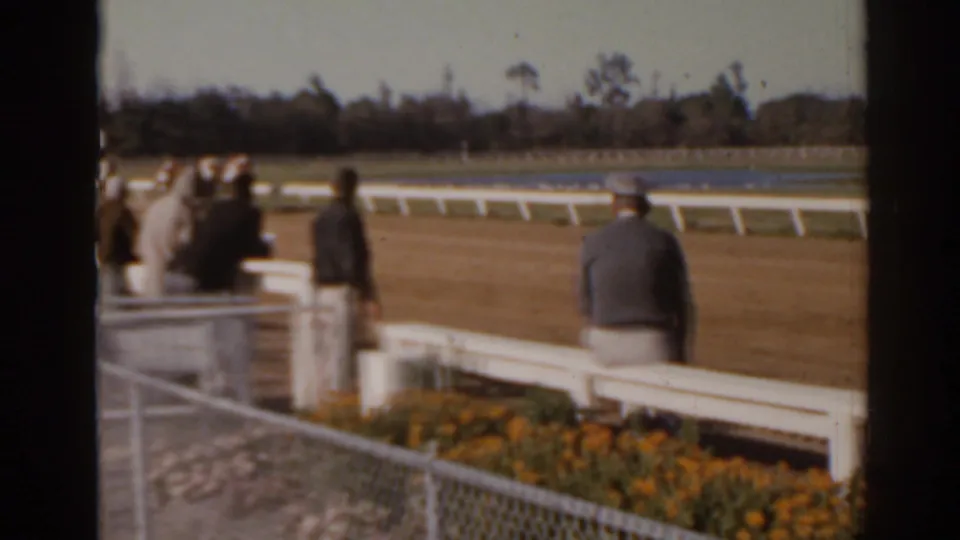 Florida 1962 home-movie frame showing spectators near a white rail as horses race in the distance.