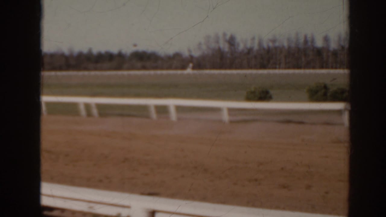Florida 1962 home-movie frame showing a dirt racetrack, white rail, and distant trees with no close people visible.