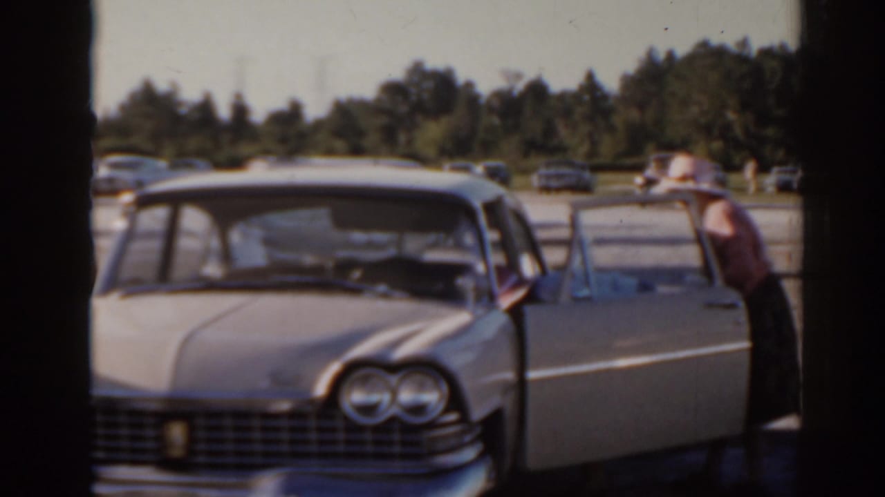 Light-colored vintage sedan parked near adults and other cars.