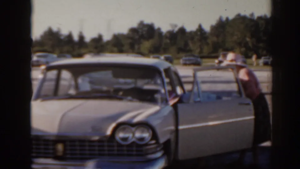 Florida 1962 frame with a light-colored 1960s sedan, open door, adults nearby, and a racetrack parking area behind it.