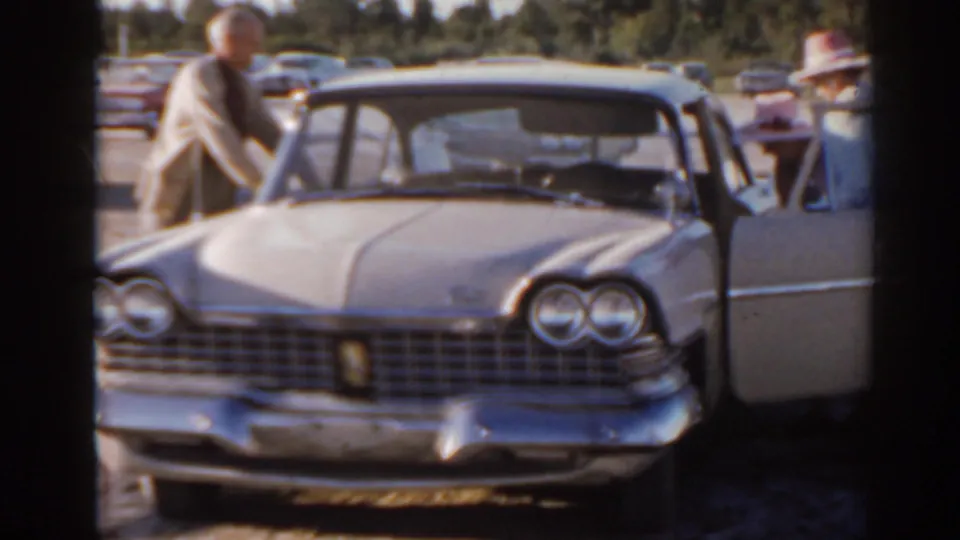 Florida 1962 frame showing adults around a light-colored 1960s sedan, including a man leaning near the hood and an open passenger door.