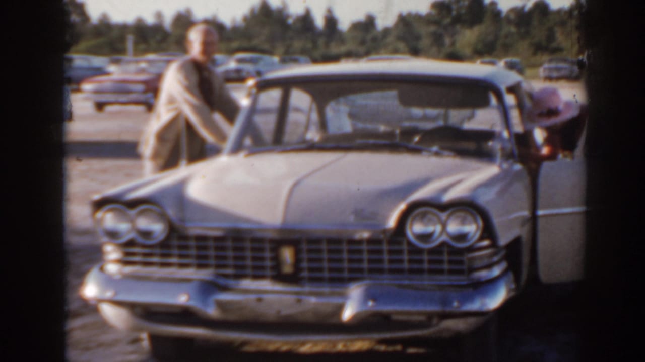 Man stands beside a light-colored classic sedan with chrome grille in a parking area.