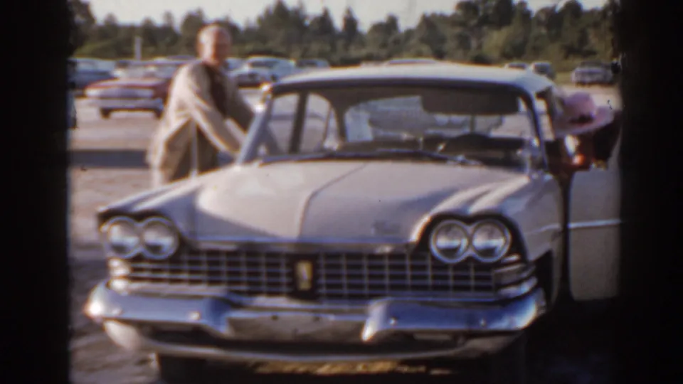 1962 Florida home-movie frame of a man by a light-colored classic sedan with quad headlights and parked cars behind it.