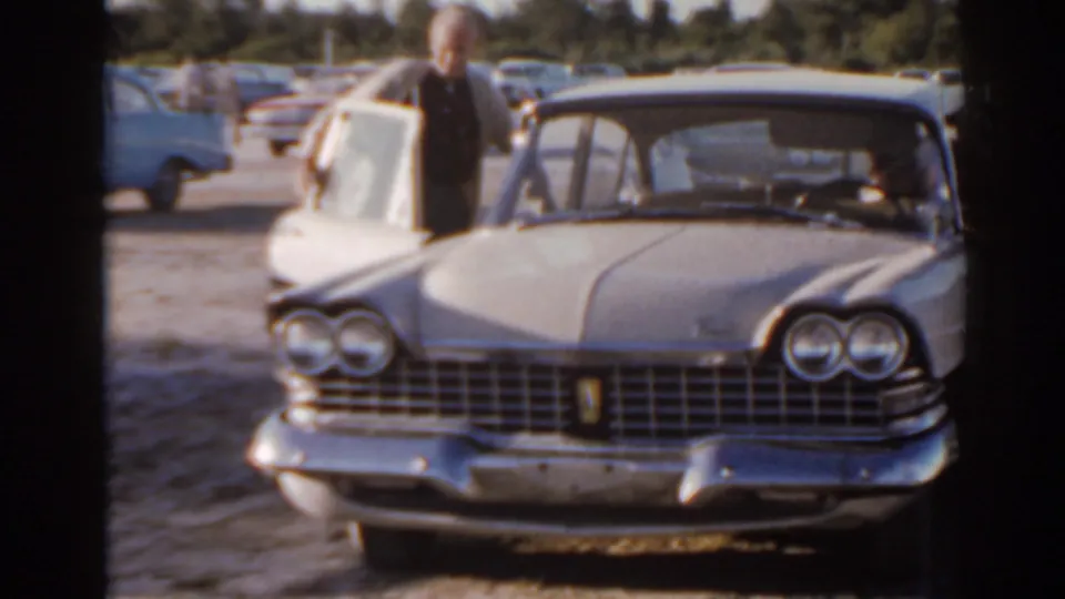 1962 Florida frame showing the front of a chrome-trimmed sedan as a man stands near an open door in sunlight.