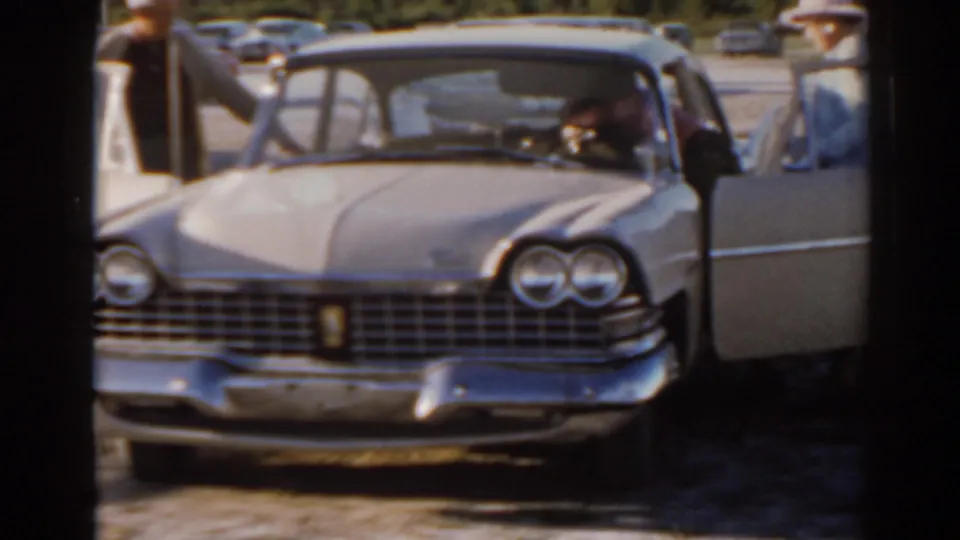 1962 Florida home-movie frame with a classic sedan, open door, and people gathered beside the car in a parking area.