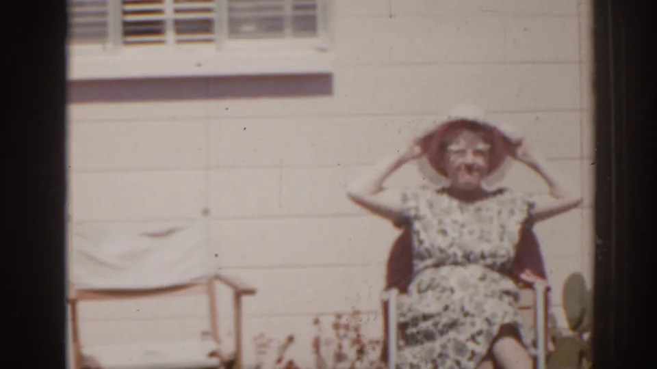 1962 Florida home-movie frame of a seated woman in a patterned dress adjusting her wide-brimmed hat outside a house wall.