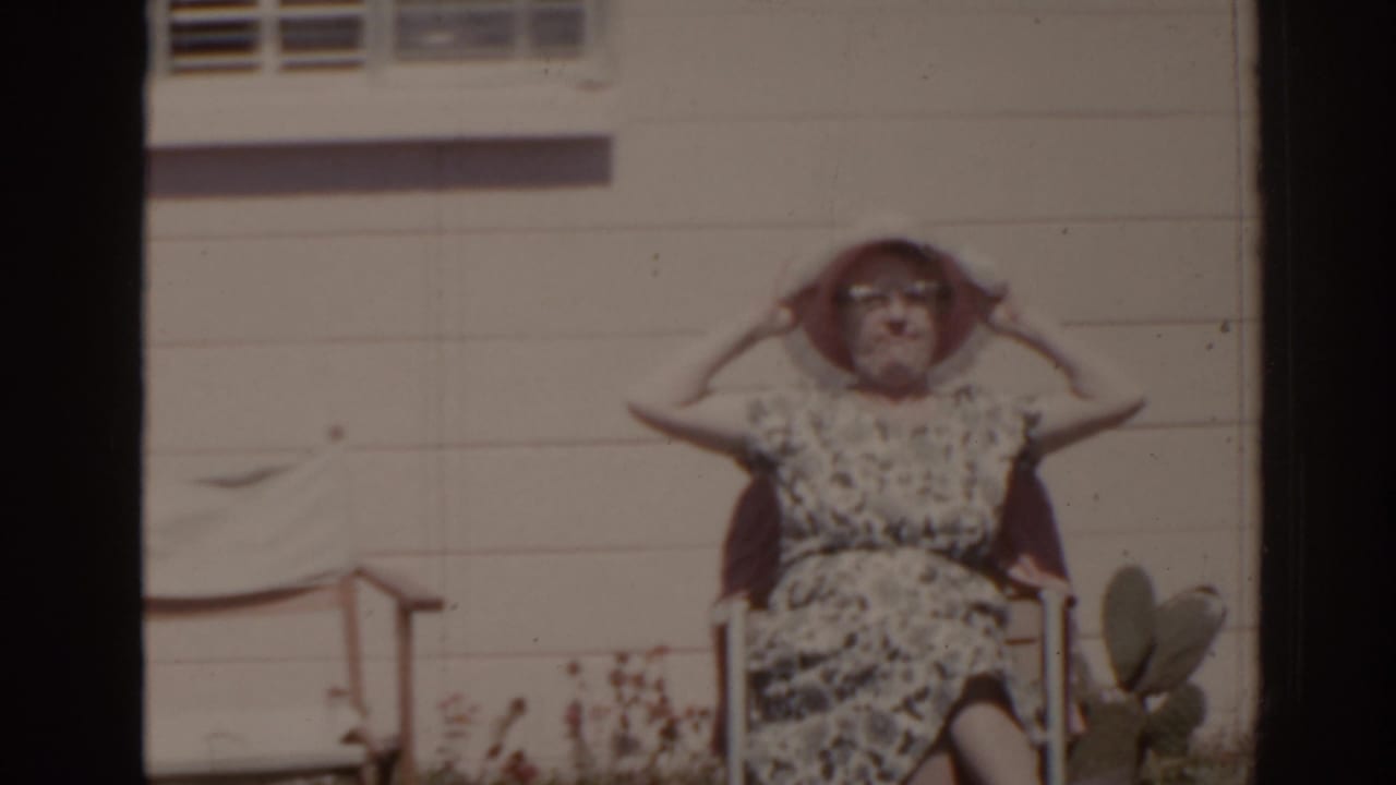 In 1962 Florida home-movie footage, a woman in a white sunhat and patterned dress sits outside near house siding and yard plants.