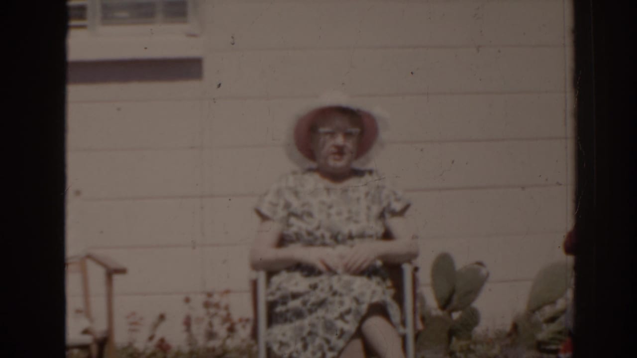 Woman in a sunhat sits with her hands folded outside a house.
