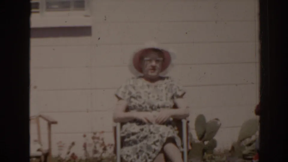 A woman in a white sunhat and patterned dress sits calmly in a chair outside a Florida house in 1962 home-movie footage.