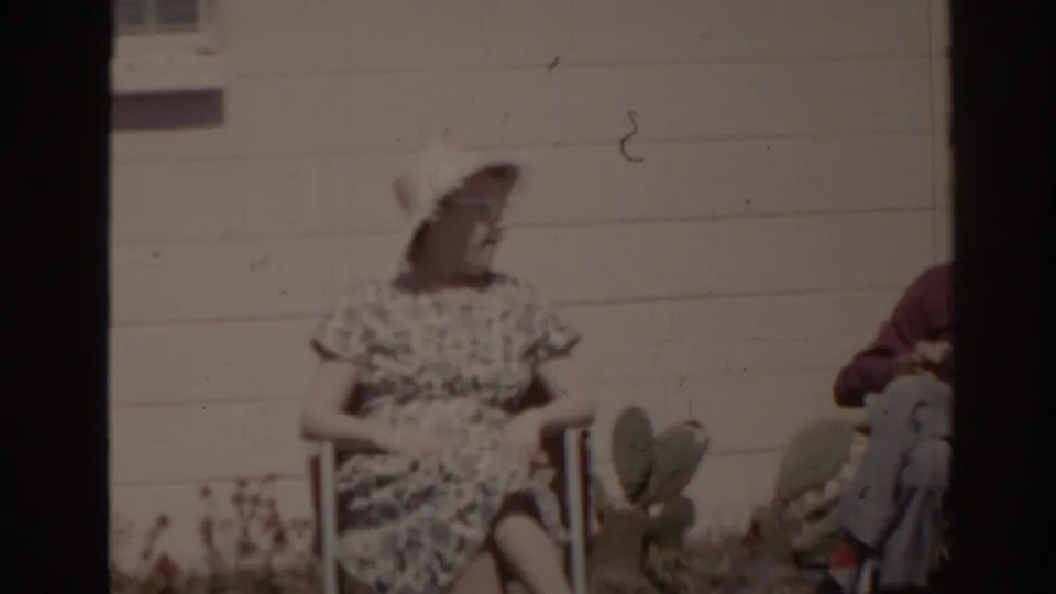 The woman turns in her chair as a man appears seated beside her, with cactus pads and house siding visible in the yard.