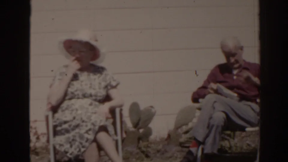 A woman in a sunhat and a man sit in outdoor chairs near cactus pads beside a house in 1962 Florida home-movie footage.
