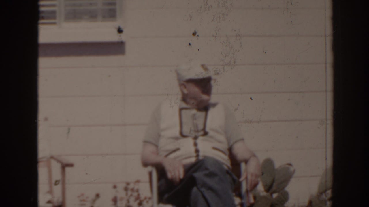 Older man in a cap seated outside against a white house wall.