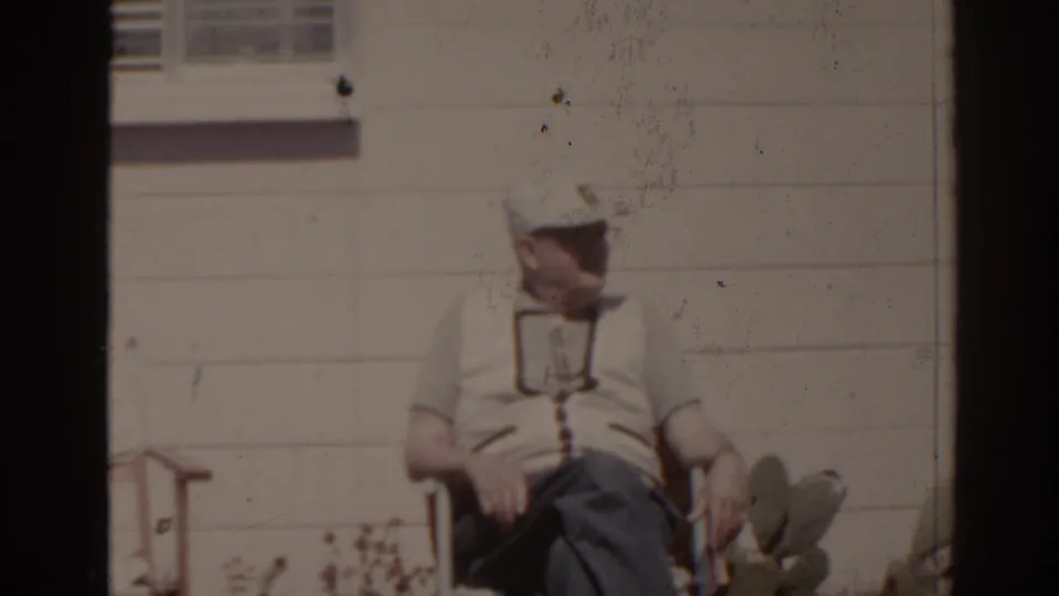 1962 Florida 8mm frame showing an older man in a light shirt and cap seated in a folding chair beside cactus plants.
