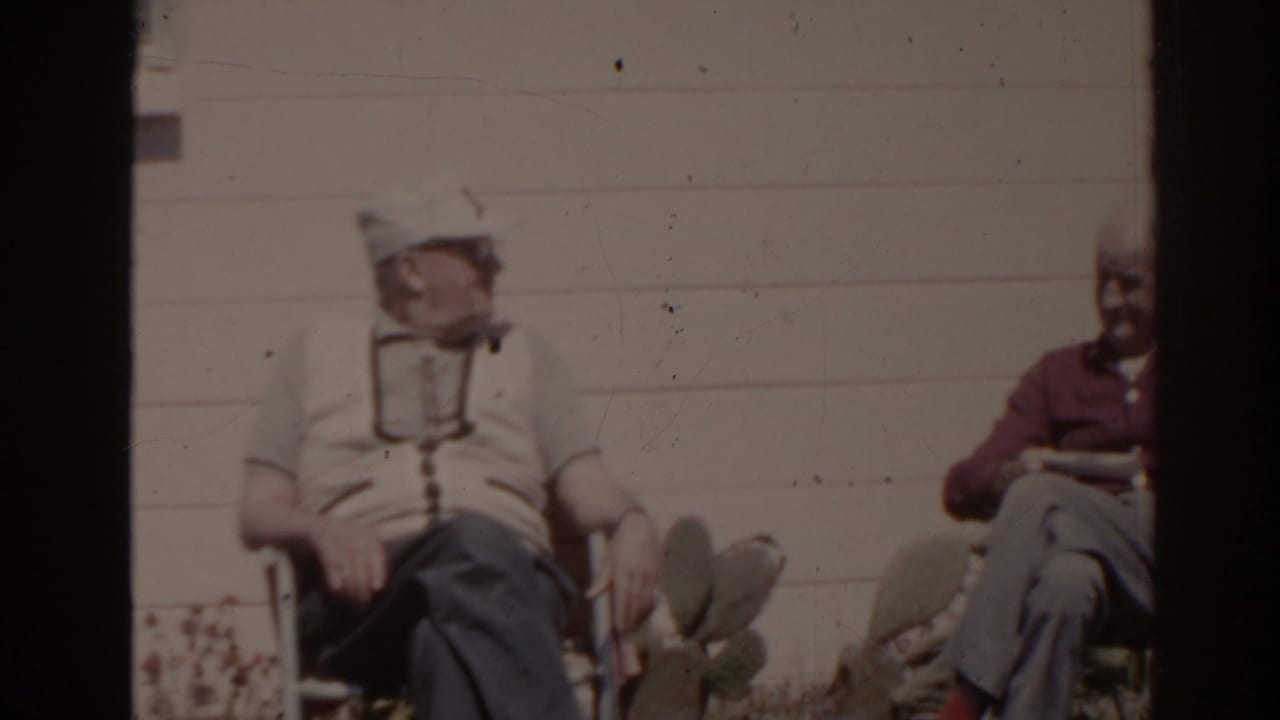 Two older men seated outdoors beside a white house wall.