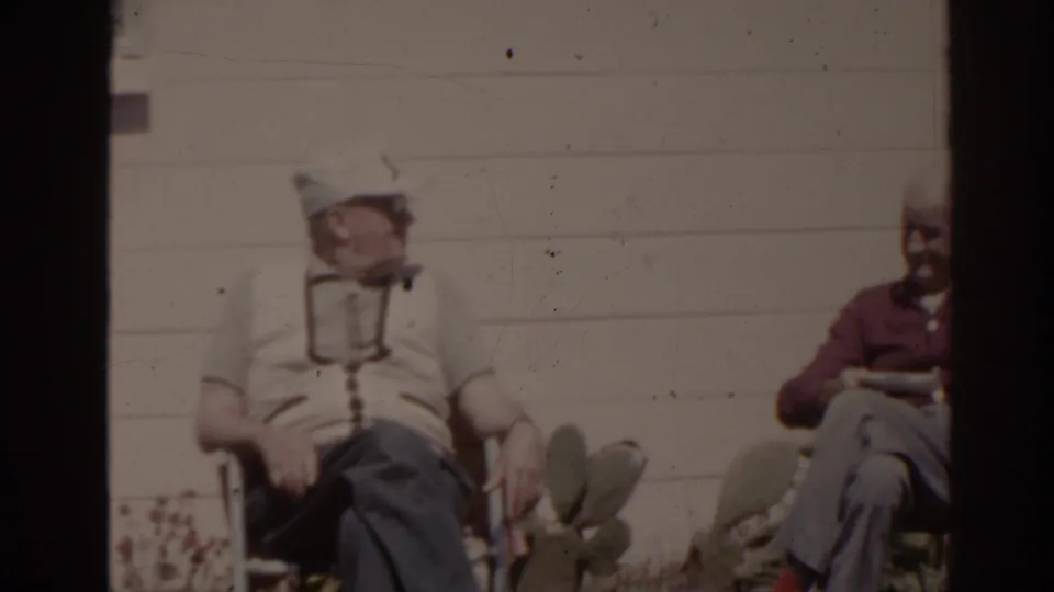 1962 Florida home-movie frame with one older man in a cap and another in a maroon shirt seated outside near cactus plants.