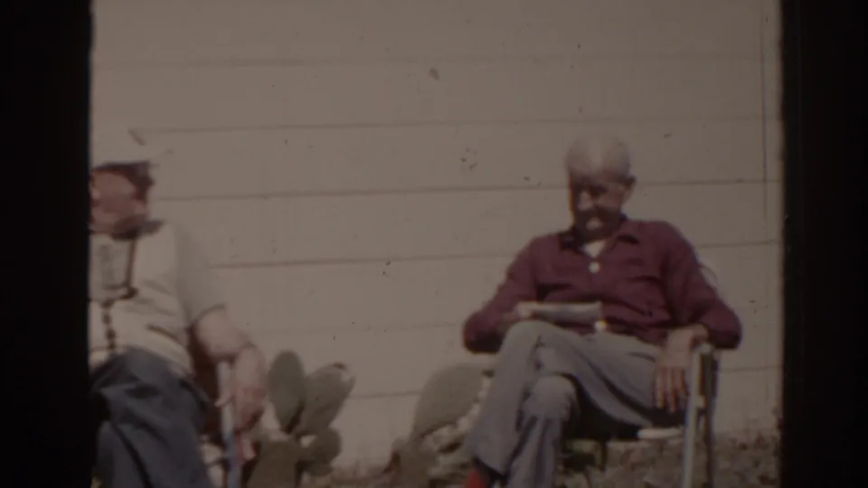 1962 Florida frame showing an older man in a maroon shirt seated outside while another seated man is partly cropped at the edge.