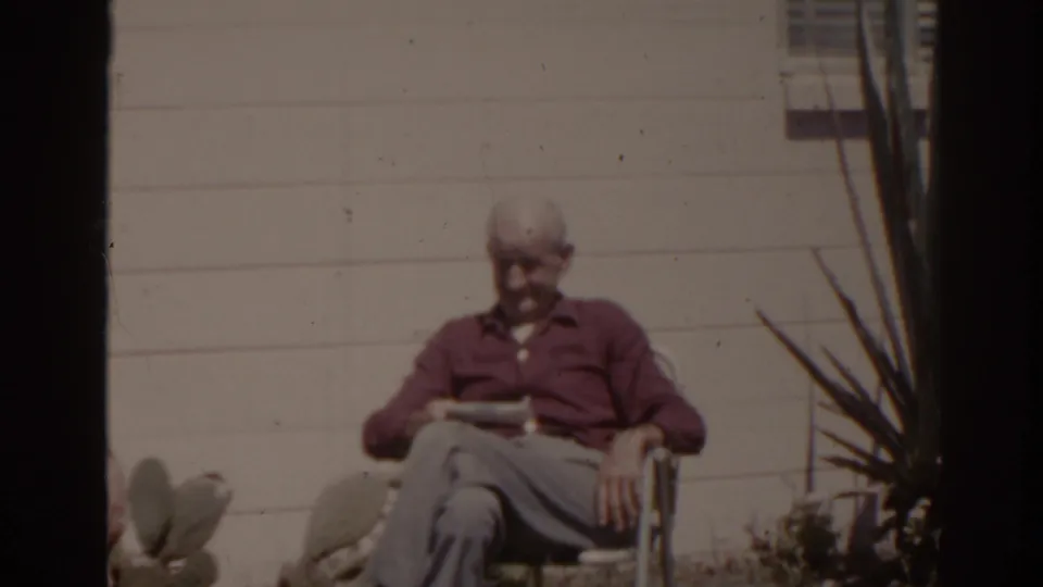 1962 Florida 8mm frame of an older man in a maroon shirt seated in a folding chair against a white house wall.