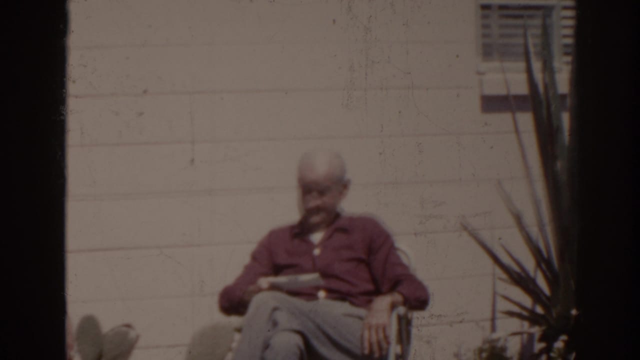 Older man in a red shirt sits outdoors reading a newspaper beside a white wall and spiky plant.