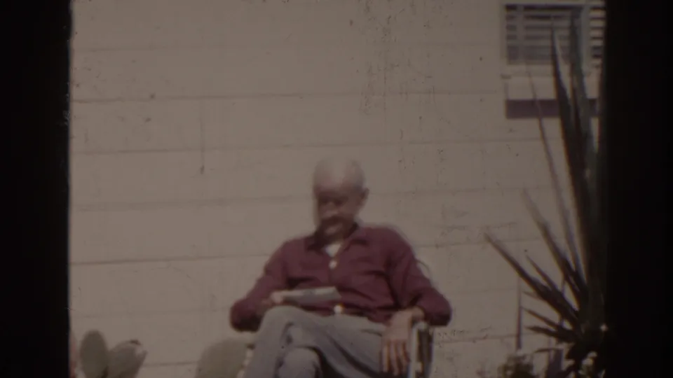 1962 Florida home-movie frame of an older man seated in a garden chair, reading a newspaper near a white exterior wall and yucca-like plant.