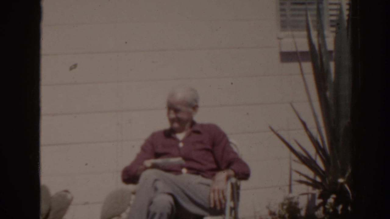 1962 Florida outdoor home-movie frame showing an older man in a red shirt reading in a chair near a shuttered window and tall plant.
