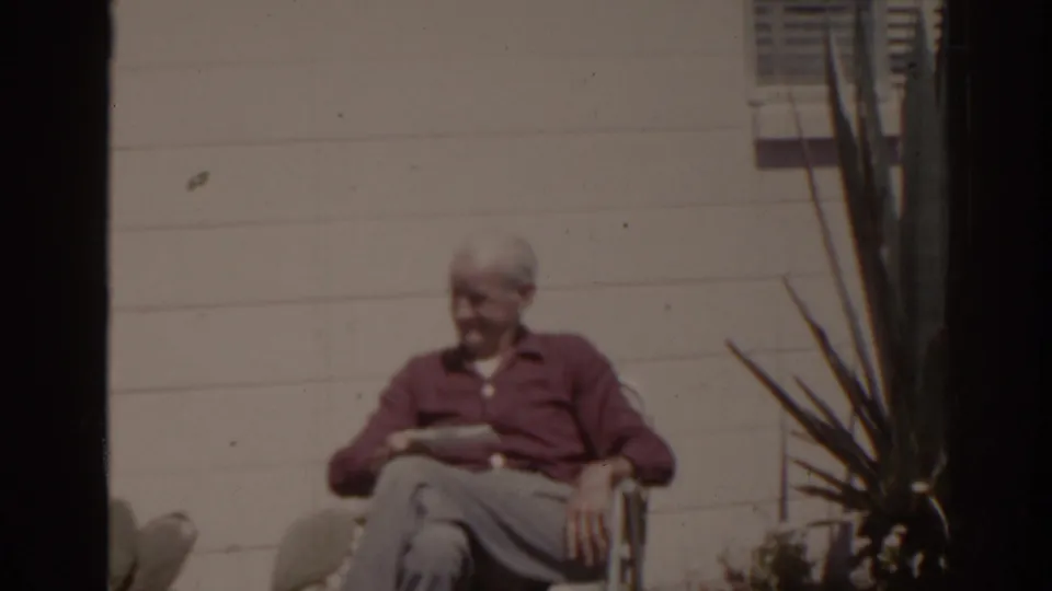 1962 Florida outdoor home-movie frame showing an older man in a red shirt reading in a chair near a shuttered window and tall plant.