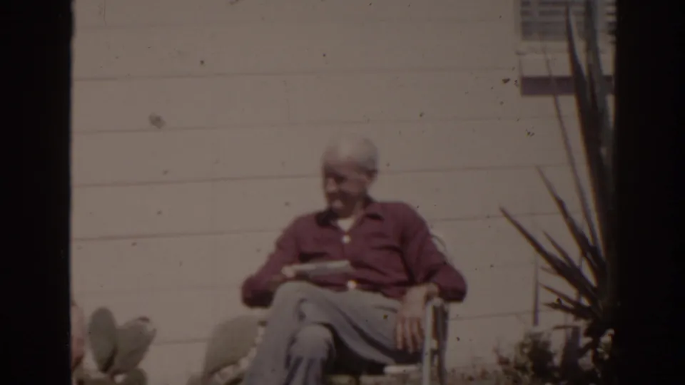 1962 Florida home-movie frame of an older man seated cross-legged outdoors, holding a newspaper beside sunlit wall siding and plants.