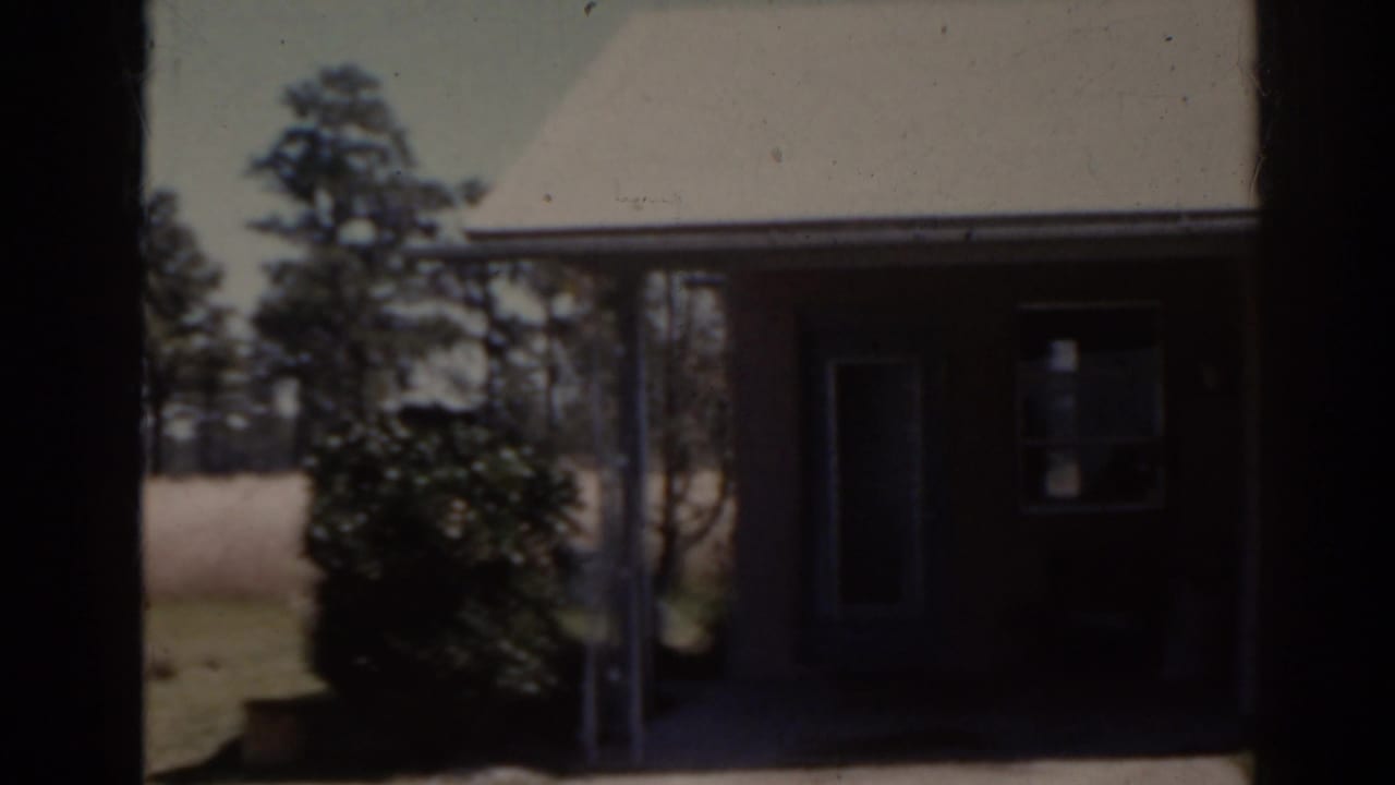 Covered porch and doorway of a single-story house with shrubs and lawn.