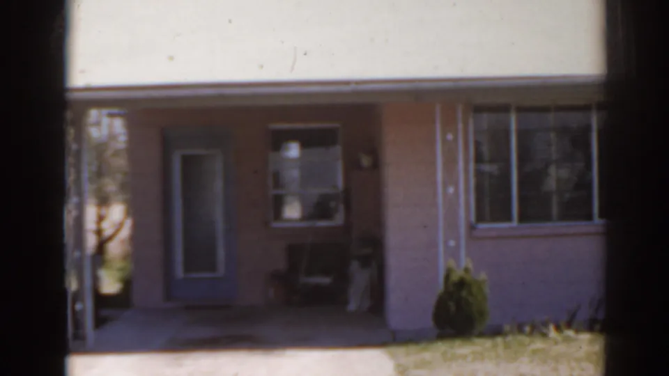 Pink single-story Florida house exterior with a blue door, wide window, small shrubs, and lawn in soft 8mm footage.